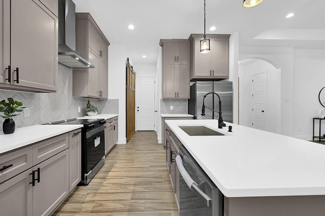 kitchen featuring sink, hanging light fixtures, stainless steel appliances, wall chimney range hood, and a center island with sink