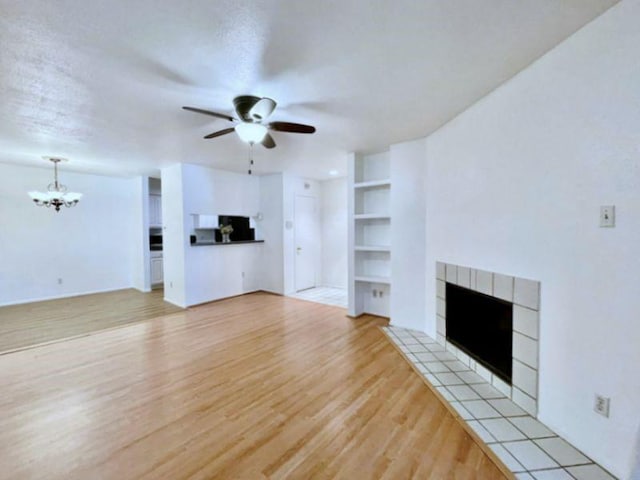 unfurnished living room featuring ceiling fan with notable chandelier, built in features, a tile fireplace, and light hardwood / wood-style flooring