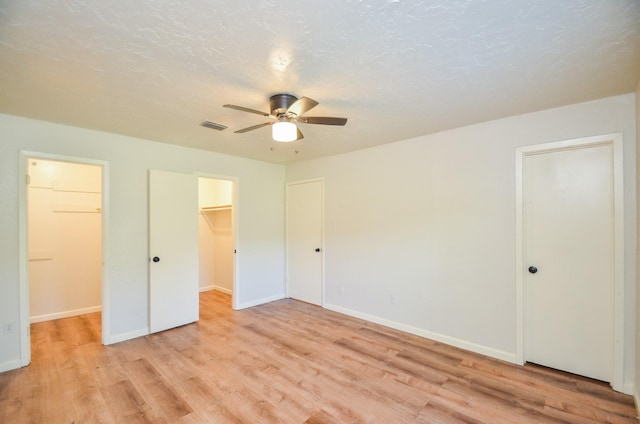 unfurnished bedroom with ceiling fan, a textured ceiling, a walk in closet, a closet, and light wood-type flooring