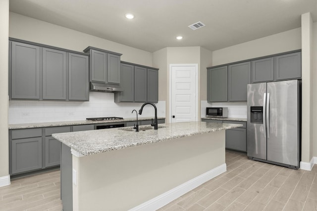 kitchen with light stone countertops, stainless steel appliances, sink, a center island with sink, and gray cabinets