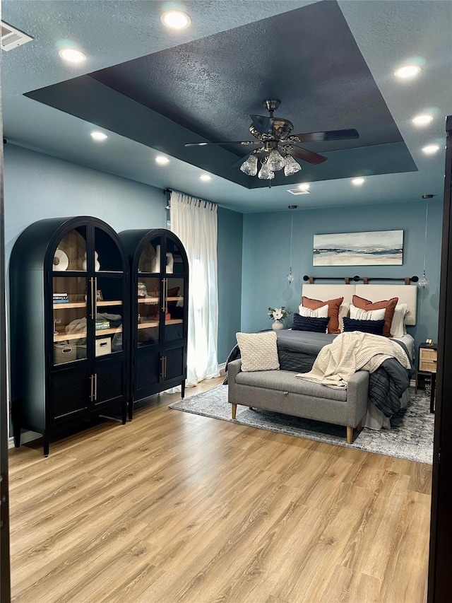 bedroom with a textured ceiling, a tray ceiling, light hardwood / wood-style flooring, and ceiling fan