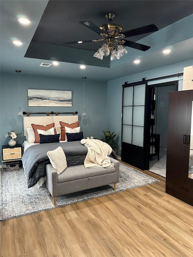 bedroom with ceiling fan, a barn door, a raised ceiling, and light wood-type flooring