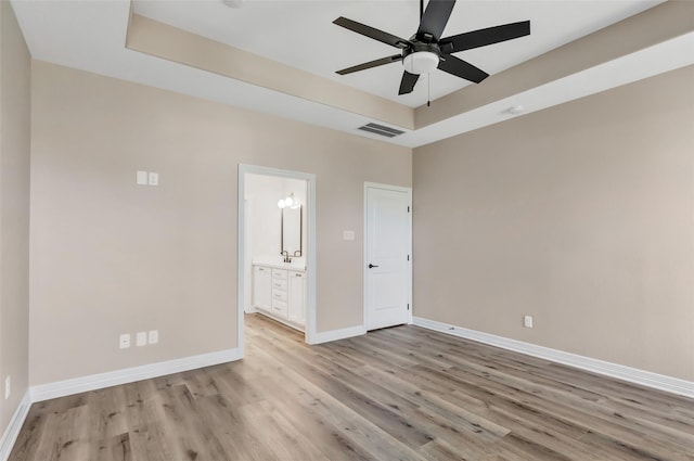 unfurnished bedroom featuring ceiling fan, light hardwood / wood-style floors, a tray ceiling, and ensuite bath