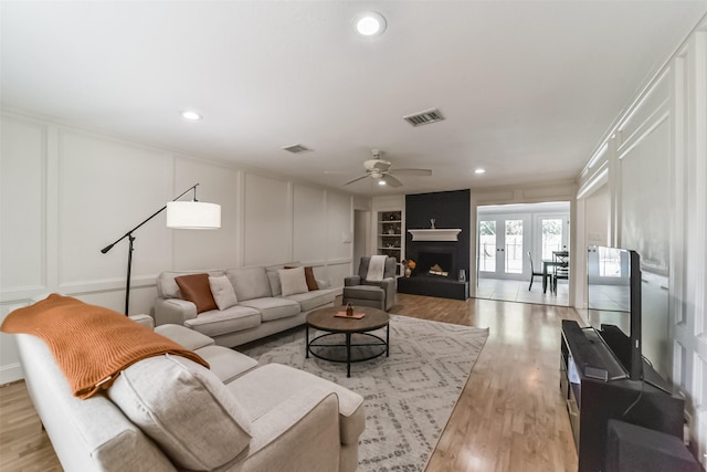 living room featuring built in shelves, crown molding, light wood-type flooring, a large fireplace, and ceiling fan