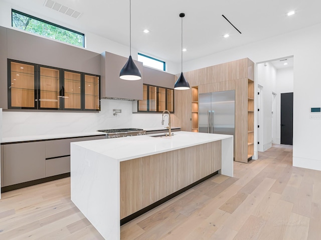 kitchen featuring sink, hanging light fixtures, backsplash, an island with sink, and light wood-type flooring