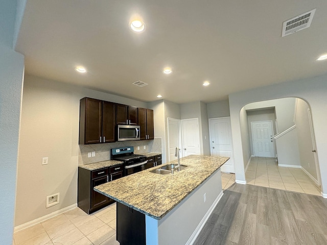 kitchen with light stone countertops, dark brown cabinets, a kitchen island with sink, sink, and black range