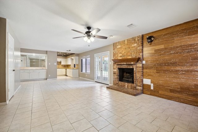 unfurnished living room featuring wood walls, french doors, a brick fireplace, ceiling fan, and light tile patterned floors