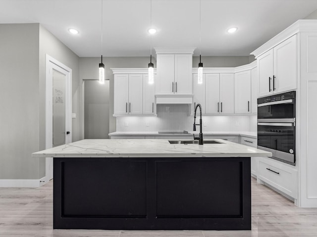 kitchen featuring light stone counters, double oven, a kitchen island with sink, pendant lighting, and white cabinets