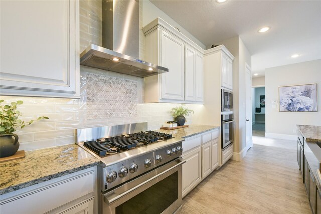 kitchen featuring wall chimney range hood, decorative backsplash, appliances with stainless steel finishes, light stone counters, and white cabinetry