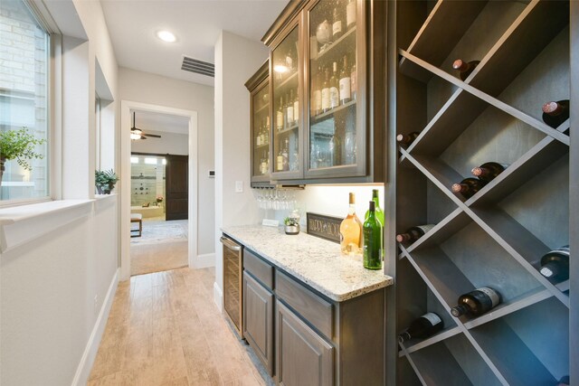 bar featuring ceiling fan, beverage cooler, light stone counters, light hardwood / wood-style flooring, and dark brown cabinets