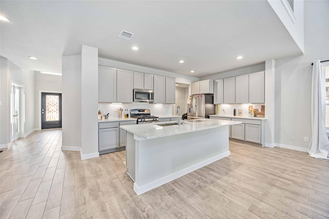 kitchen featuring gray cabinetry, light stone counters, a center island with sink, appliances with stainless steel finishes, and light hardwood / wood-style floors