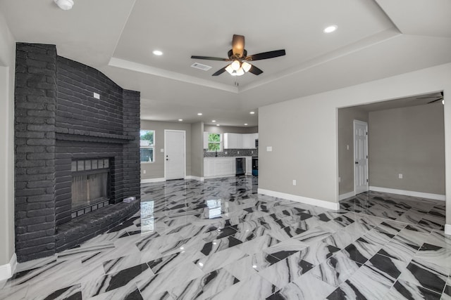 unfurnished living room featuring sink, a fireplace, ceiling fan, and a tray ceiling