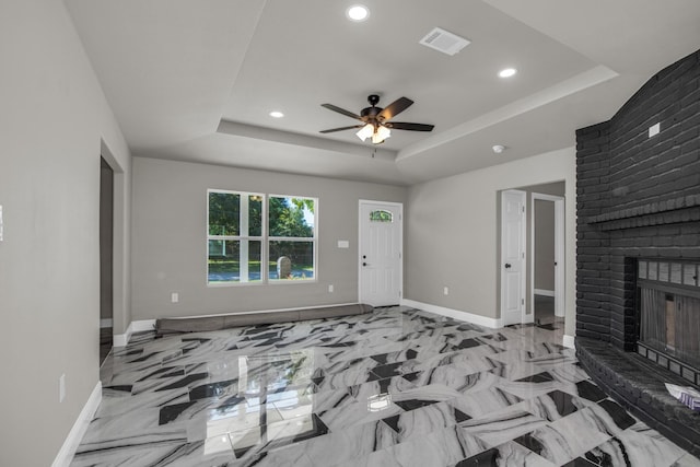 unfurnished living room with ceiling fan, a tray ceiling, and a brick fireplace