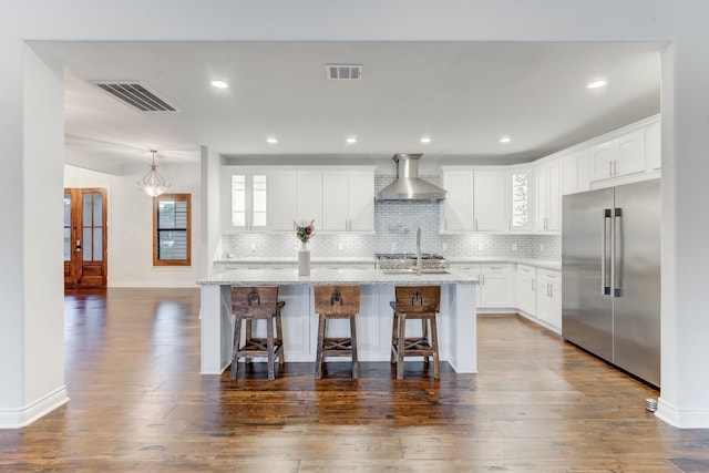 kitchen featuring white cabinets, wall chimney exhaust hood, stainless steel built in refrigerator, a kitchen island with sink, and a breakfast bar area