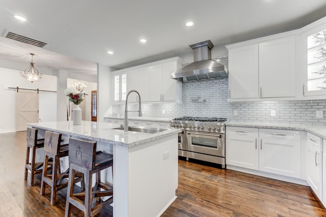 kitchen with a barn door, decorative backsplash, range with two ovens, wall chimney range hood, and white cabinets