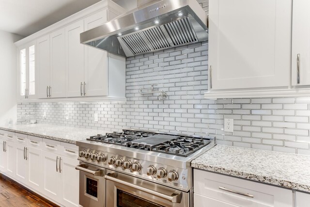 kitchen featuring decorative backsplash, white cabinetry, range with two ovens, wall chimney exhaust hood, and light stone counters