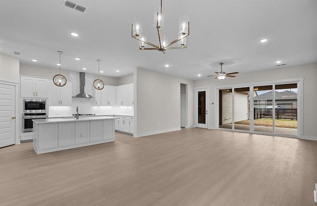 kitchen featuring appliances with stainless steel finishes, wall chimney range hood, pendant lighting, white cabinets, and an island with sink