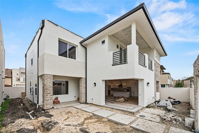 rear view of house featuring stone siding, fence, a balcony, and stucco siding