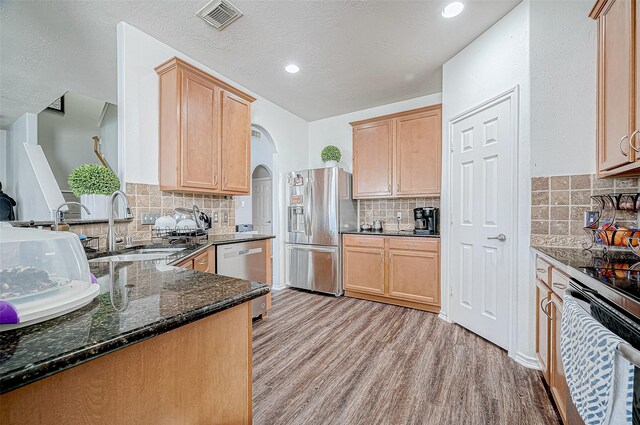 kitchen with kitchen peninsula, appliances with stainless steel finishes, tasteful backsplash, light hardwood / wood-style flooring, and dark stone countertops