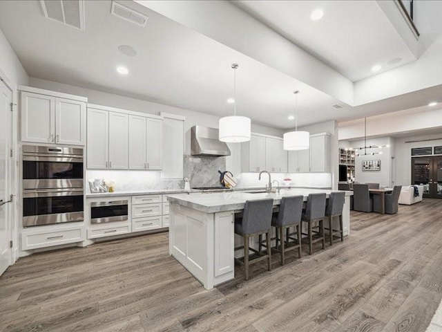 kitchen featuring white cabinets, a center island with sink, wall chimney range hood, appliances with stainless steel finishes, and light hardwood / wood-style floors