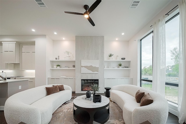 living room with ceiling fan, a large fireplace, built in shelves, and dark hardwood / wood-style flooring