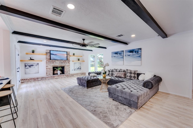 living room with ceiling fan, a brick fireplace, light hardwood / wood-style flooring, beamed ceiling, and a textured ceiling