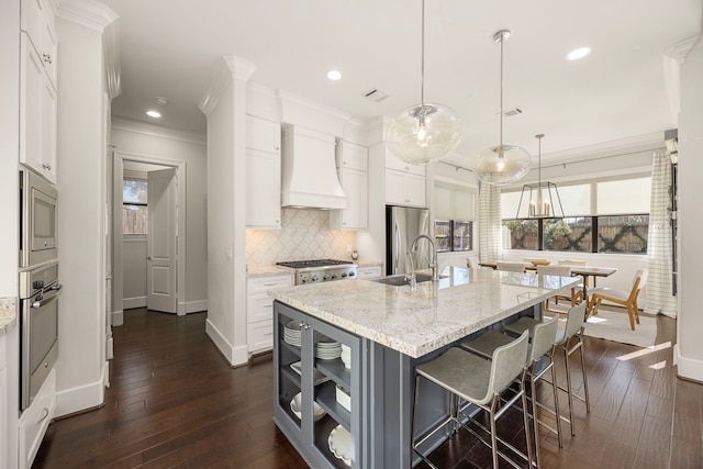 kitchen with white cabinets, an island with sink, decorative light fixtures, custom range hood, and stainless steel appliances