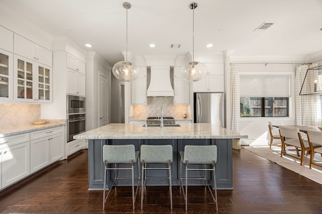 kitchen featuring pendant lighting, premium range hood, white cabinets, an island with sink, and stainless steel appliances