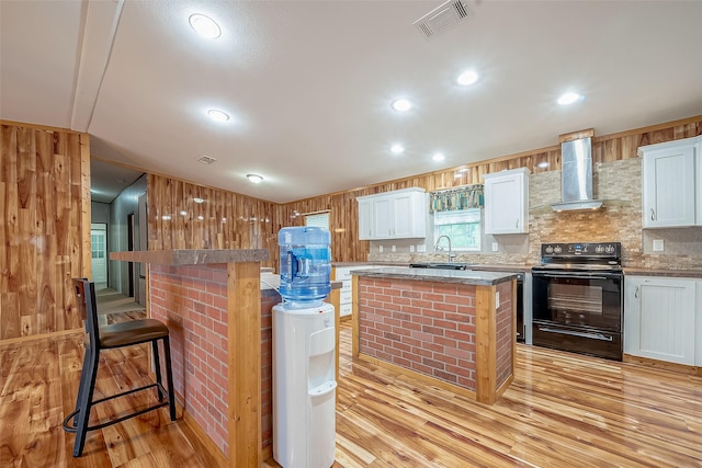 kitchen featuring a center island, black electric range oven, white cabinets, wall chimney range hood, and a kitchen bar