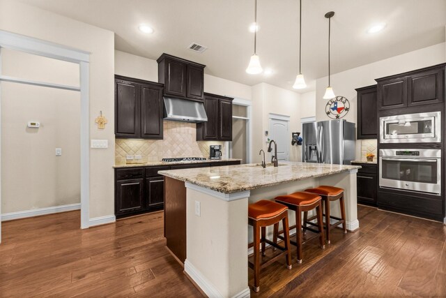 kitchen featuring a kitchen bar, appliances with stainless steel finishes, backsplash, a kitchen island with sink, and hanging light fixtures