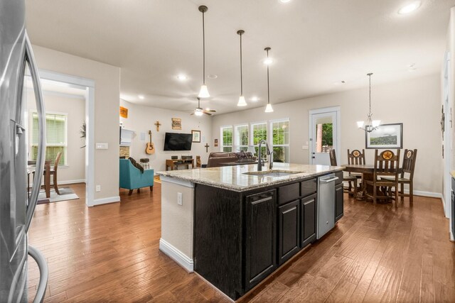 kitchen with appliances with stainless steel finishes, light stone counters, ceiling fan with notable chandelier, sink, and a center island with sink