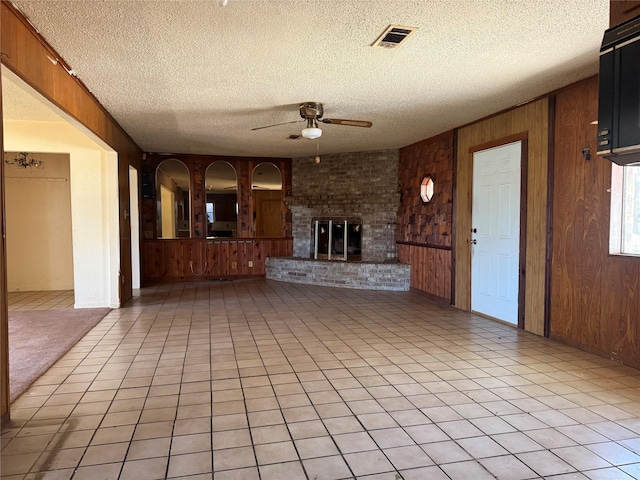 unfurnished living room with wood walls, ceiling fan, a textured ceiling, and a brick fireplace