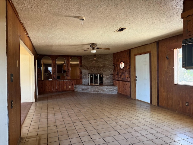 unfurnished living room featuring wooden walls, a brick fireplace, ceiling fan, light tile patterned floors, and a textured ceiling