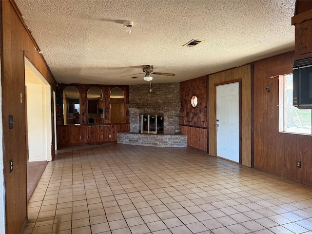 unfurnished living room featuring ceiling fan, wood walls, a textured ceiling, and a brick fireplace