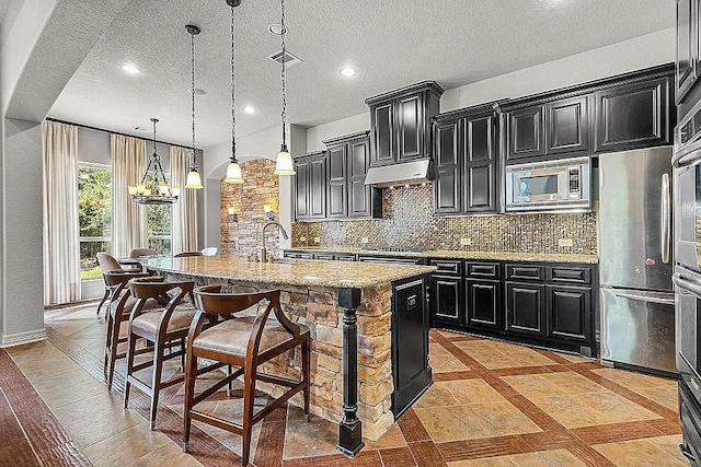 kitchen featuring light stone countertops, decorative light fixtures, a kitchen bar, a kitchen island with sink, and appliances with stainless steel finishes