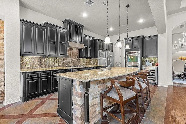 kitchen featuring wine cooler, an island with sink, light stone counters, a breakfast bar area, and stainless steel appliances