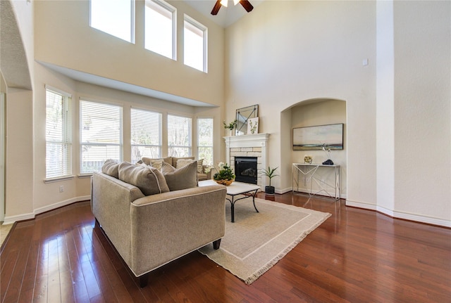 living room featuring a high ceiling, a stone fireplace, ceiling fan, and dark wood-type flooring