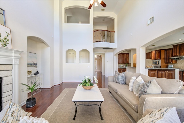 living room with ceiling fan, a towering ceiling, a fireplace, and dark wood-type flooring