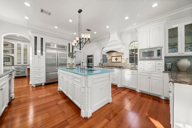 kitchen featuring backsplash, premium range hood, hanging light fixtures, built in appliances, and an island with sink