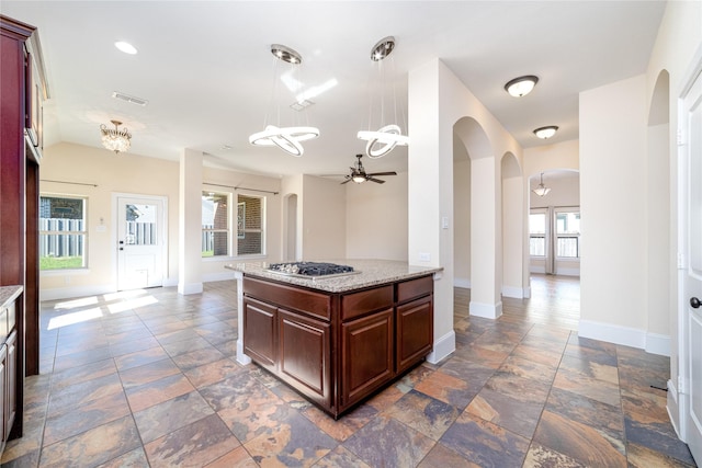 kitchen with light stone counters, ceiling fan with notable chandelier, pendant lighting, a center island, and stainless steel gas stovetop