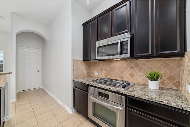 kitchen with light tile patterned floors, appliances with stainless steel finishes, dark brown cabinetry, light stone countertops, and decorative backsplash