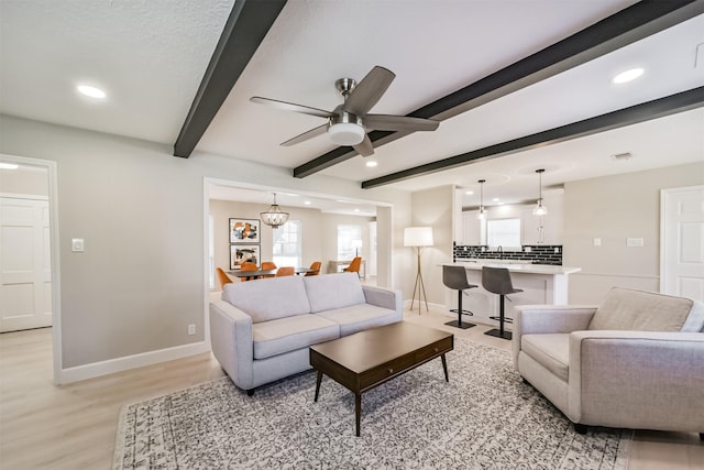 living room featuring beamed ceiling, ceiling fan with notable chandelier, and light wood-type flooring