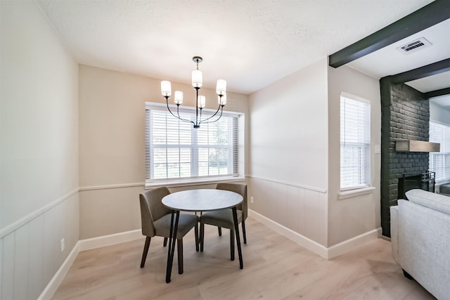 dining area with a brick fireplace, light hardwood / wood-style flooring, a textured ceiling, beamed ceiling, and a notable chandelier