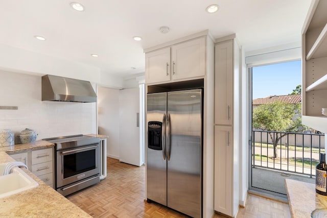 kitchen with stainless steel appliances, sink, white cabinets, wall chimney exhaust hood, and light parquet flooring