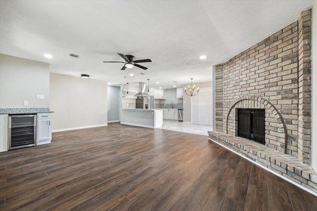 unfurnished living room featuring ceiling fan with notable chandelier, sink, a brick fireplace, dark hardwood / wood-style flooring, and beverage cooler