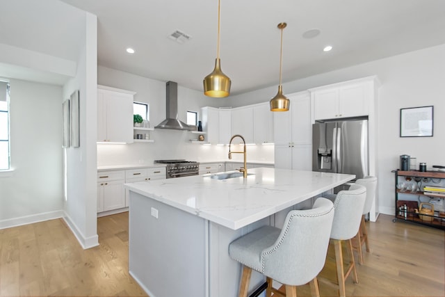 kitchen with an island with sink, sink, white cabinets, hanging light fixtures, and wall chimney range hood