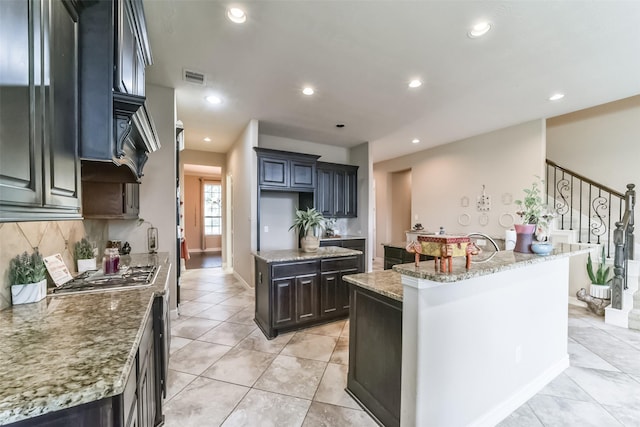 kitchen with decorative backsplash, light tile patterned floors, light stone counters, and a kitchen island with sink