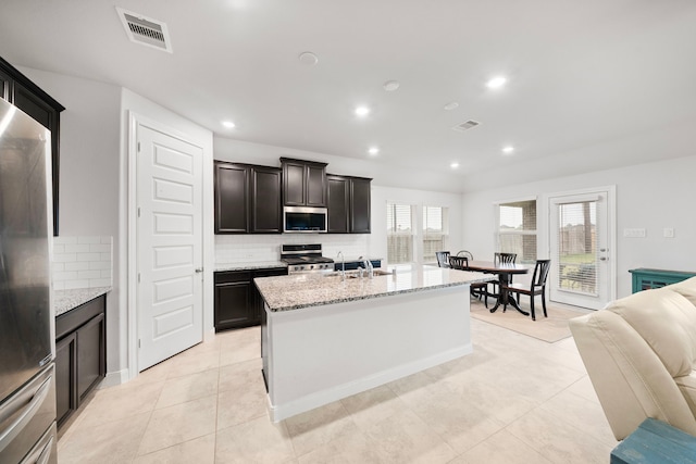 kitchen featuring appliances with stainless steel finishes, backsplash, light stone countertops, a healthy amount of sunlight, and a center island with sink