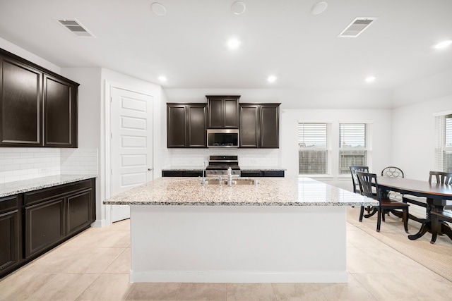 kitchen with dark brown cabinetry, light tile patterned floors, an island with sink, stainless steel appliances, and backsplash