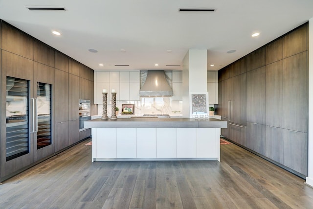 kitchen with a spacious island, wall chimney range hood, double wall oven, and light hardwood / wood-style flooring